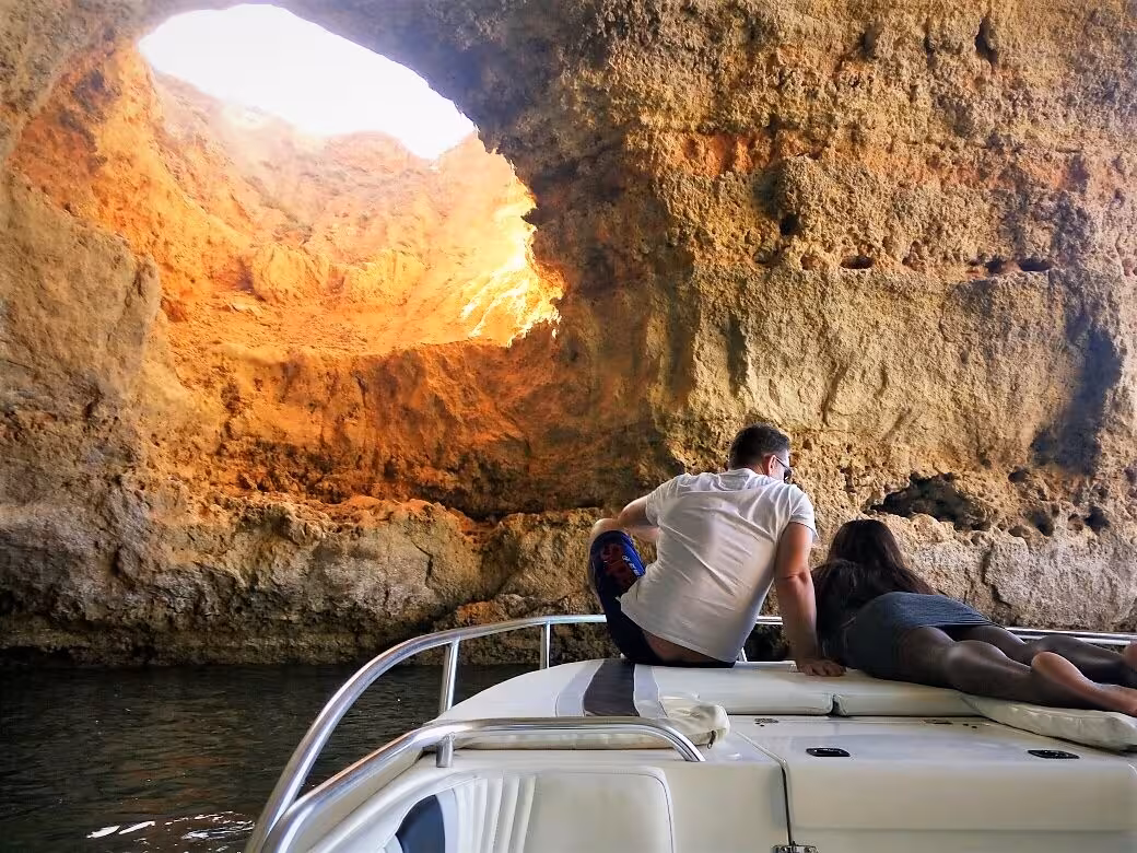 Couple relaxing on private boat tour, cruising into dramatic Algarve sea cave with golden rock walls glowing at sunset
