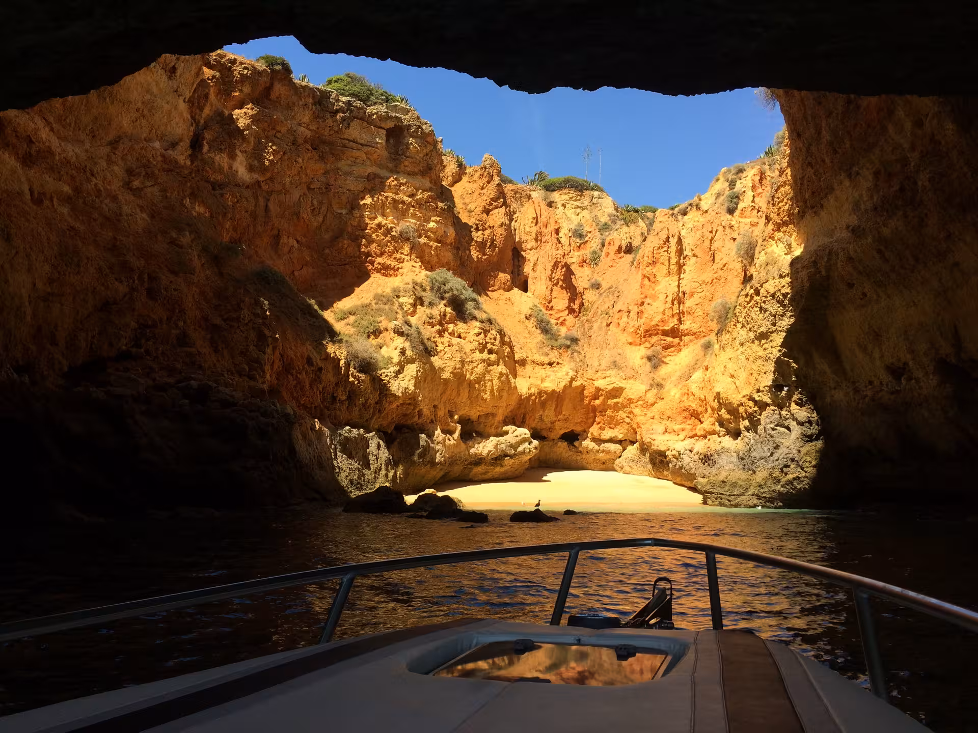 Private boat gliding into secluded Algarve sea cave, revealing hidden sandy beach and towering sunlit orange cliffs