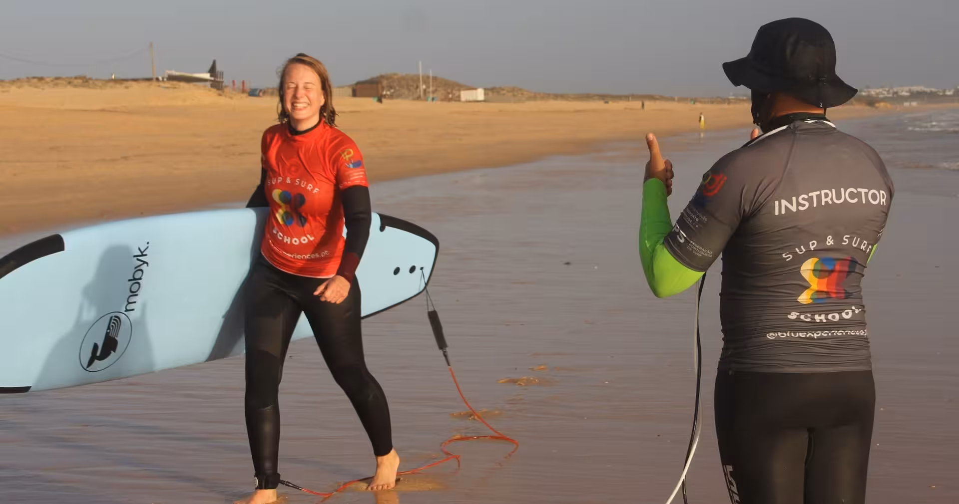 Smiling beginner carrying a soft-top surfboard as a certified surf instructor gives feedback during a private beach lesson