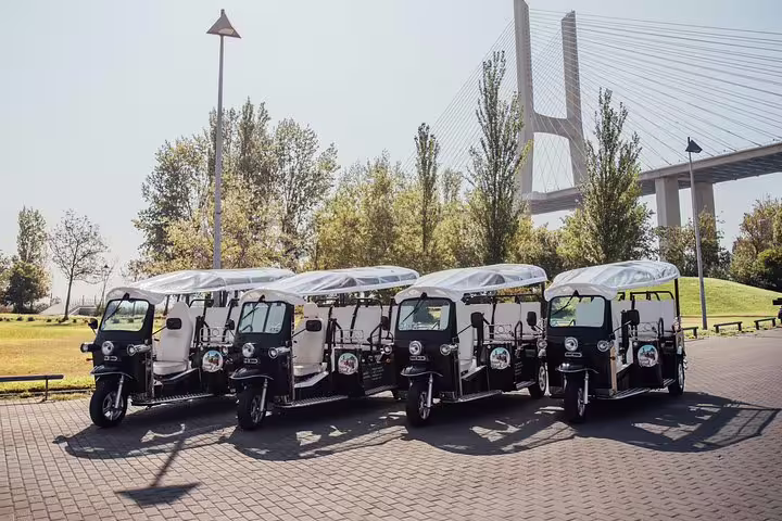 Parked tuk tuks ready for a private 4-hour Lisbon discovery tour, with the iconic Vasco da Gama Bridge in the background.