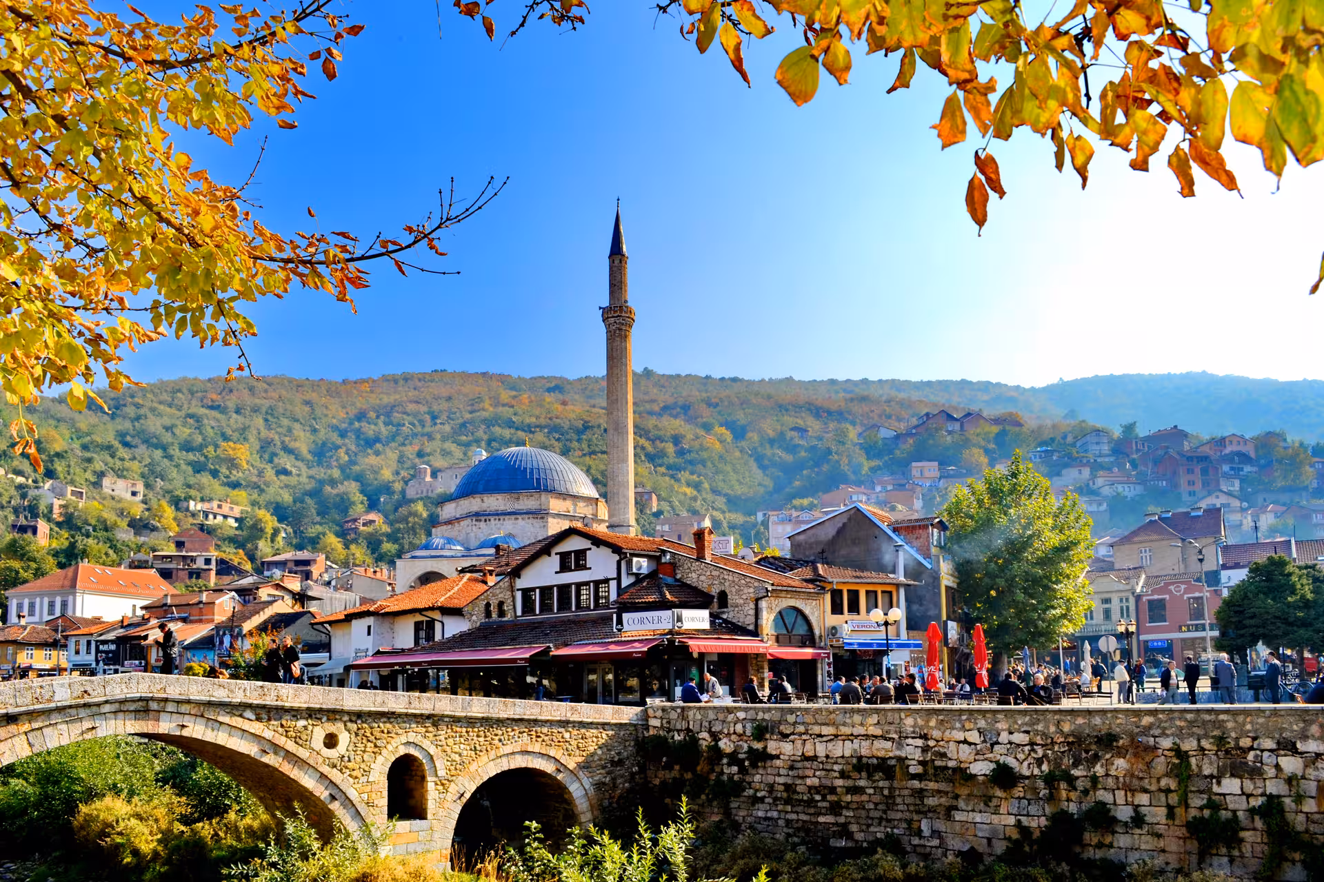 Historic stone bridge and mosque in Prizren, Kosovo surrounded by autumn foliage and hillside homes.