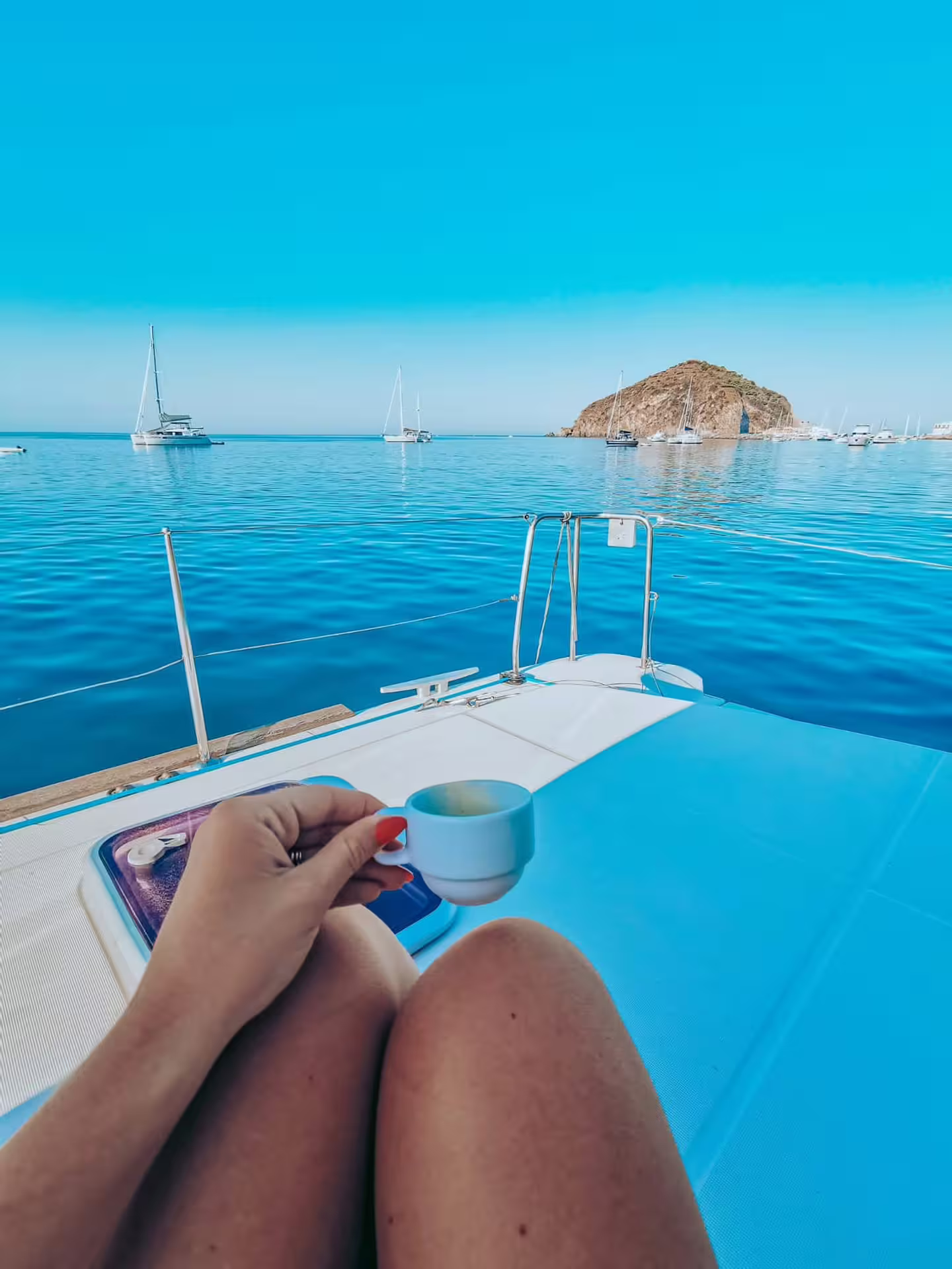 Guest sipping espresso on catamaran deck during Procida sailing tour, facing turquoise sea, anchored boats and rocky islet