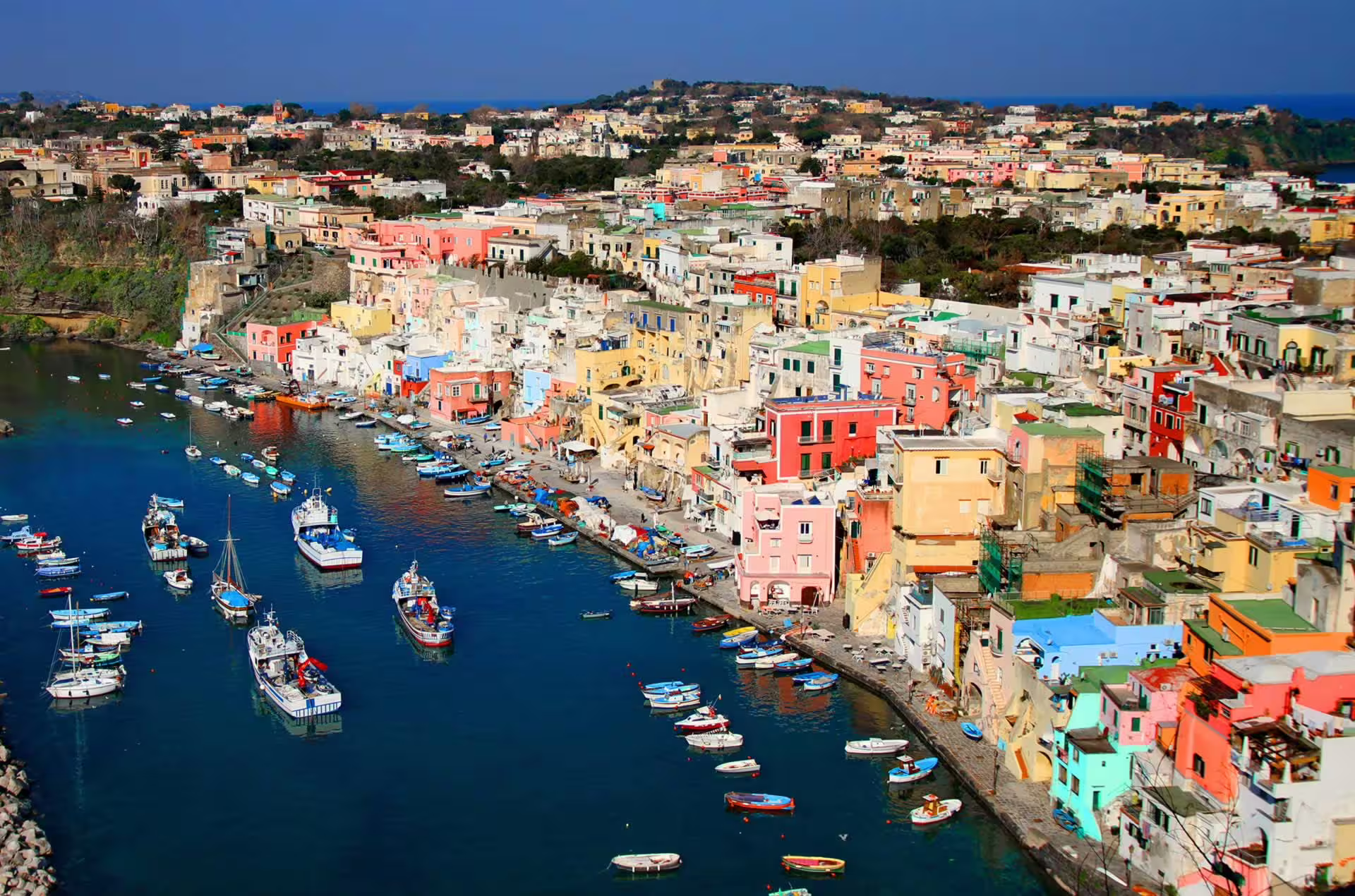 Aerial view of colorful Procida harbor with fishing boats, pastel houses and blue sea on Ischia & Procida boat trip from Naples