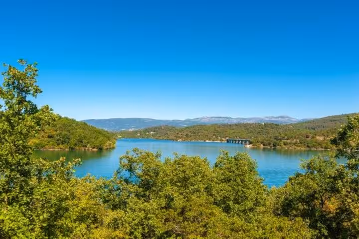 Scenic view of a tranquil lake surrounded by lush greenery in the Provencal countryside under a clear blue sky.