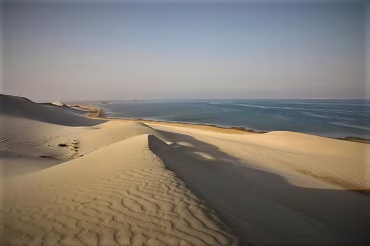 Expansive view of Qatar's golden sand dunes meeting the serene blue waters of the Inland Sea desert.