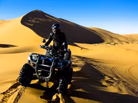 Rider on a quad bike navigating the golden sand dunes of Merzouga Erg Chebbi Desert under a clear blue sky.