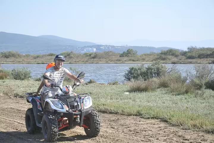 Father and child riding a quad bike along a scenic path with a lake and hills near Kusadasi, Turkey.