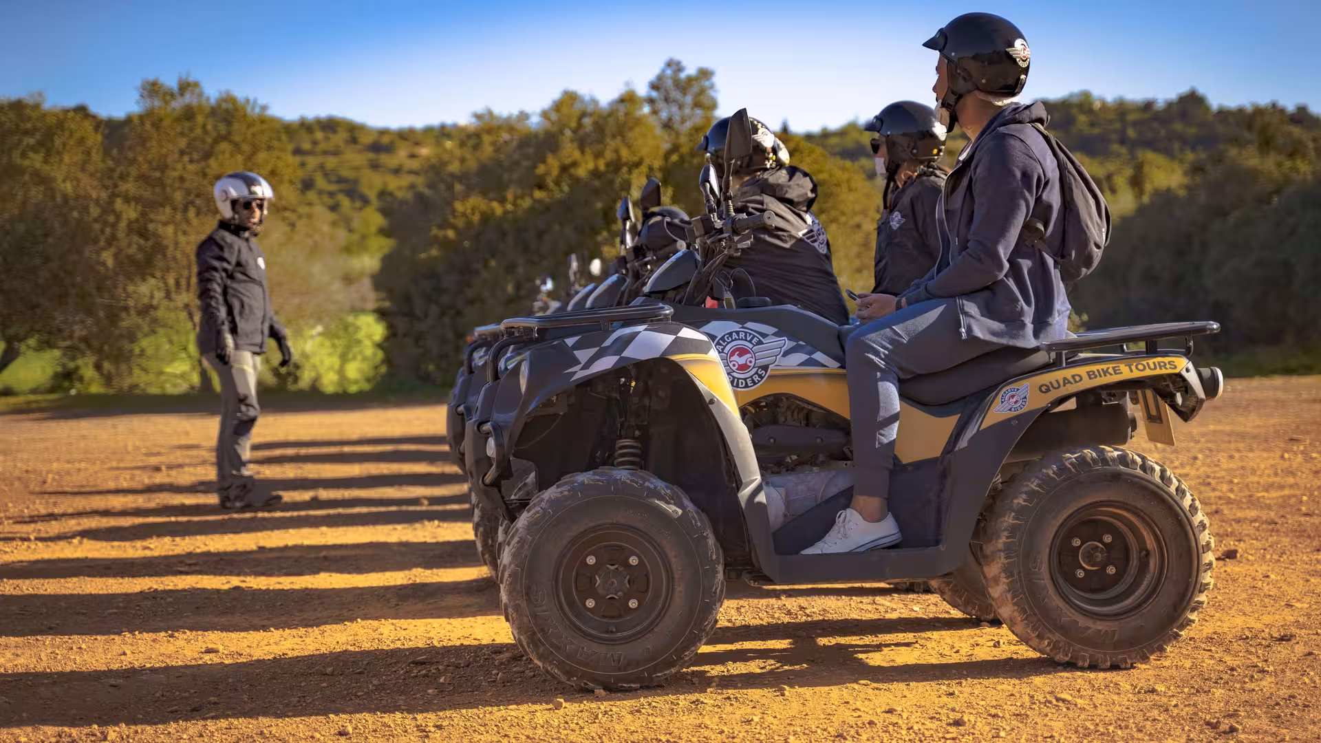 Quad tour briefing as helmeted riders sit on parked ATVs listening to their guide on a sunny dirt track in the countryside