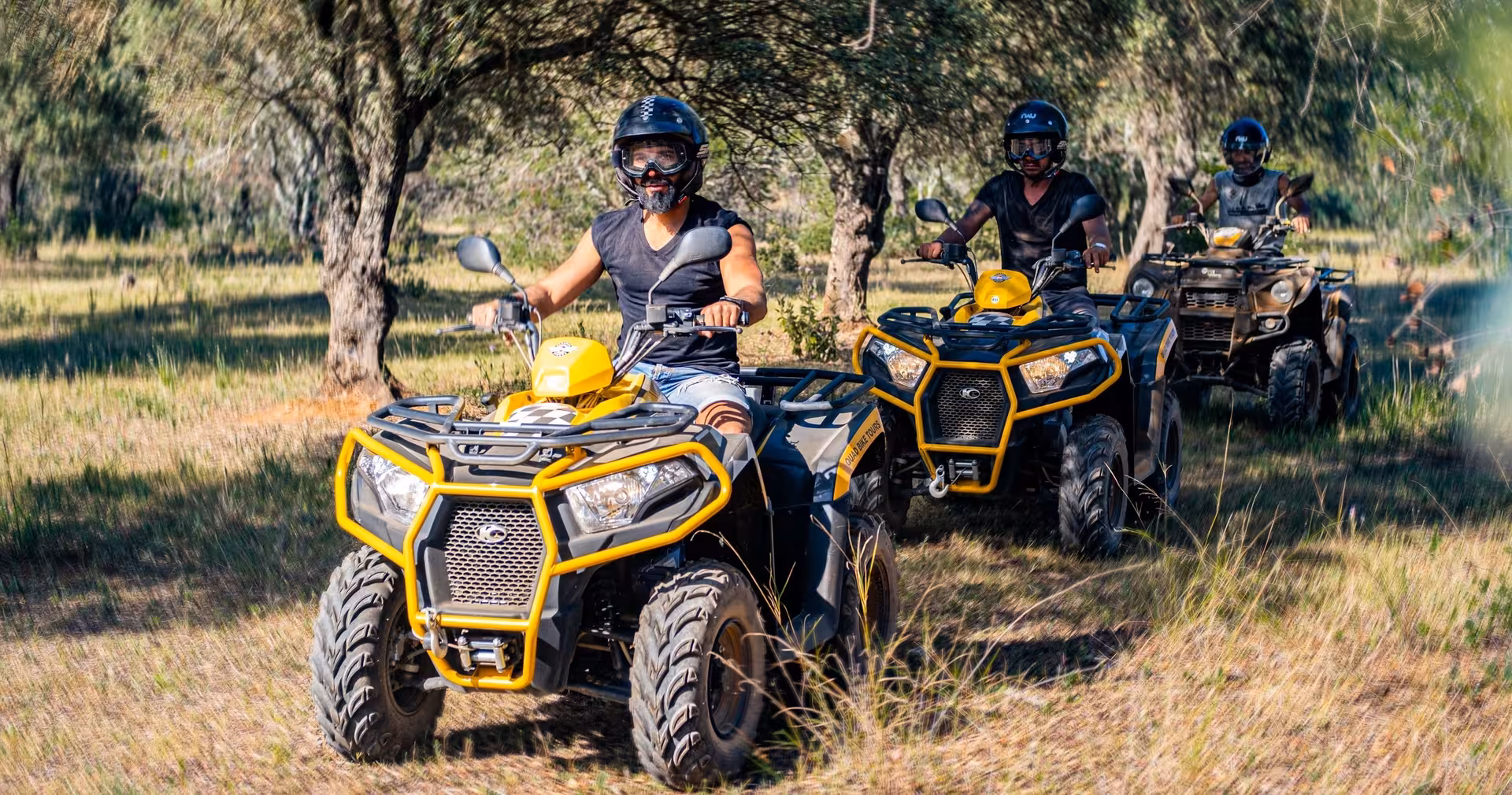 Guided quad tour adventure with riders driving yellow ATVs along a scenic forest trail through grass and olive trees