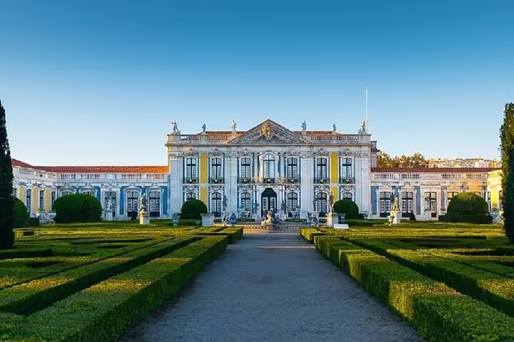 Elegant facade of Queluz Palace with manicured gardens under a clear blue sky, part of the Mafra, Ericeira, and Queluz tour.