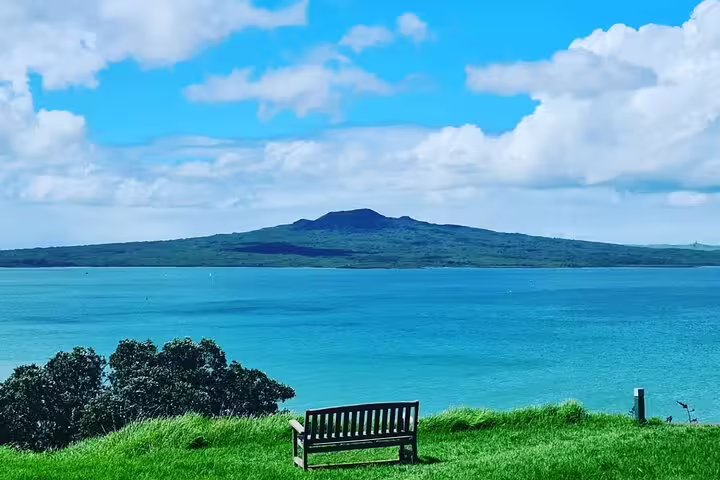 Scenic view of Rangitoto Island from a grassy hill in Auckland with a bench overlooking the blue ocean.