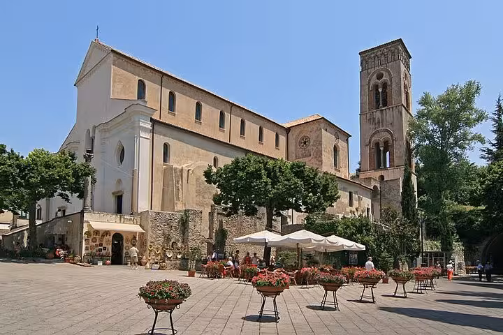 Historic cathedral in Ravello with Romanesque architecture, vibrant flower pots, and shaded outdoor seating areas.