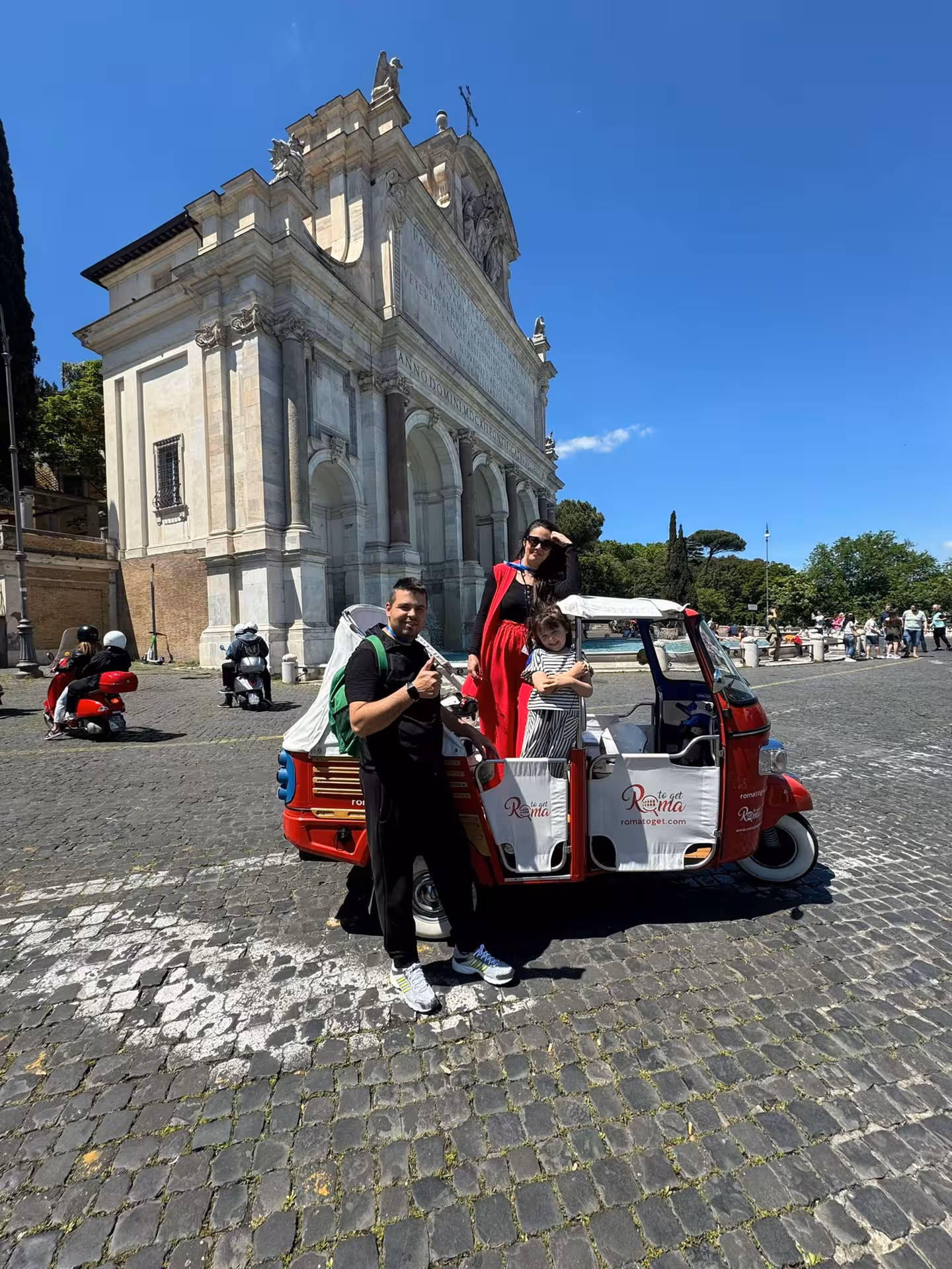 Visitors pose by a red Ape Calessino in front of Rome's historic architecture, highlighting a unique city tour.