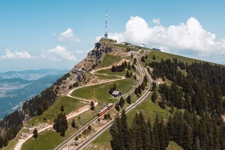 Aerial view of a red cogwheel train ascending a winding track to the summit of a Swiss mountain, surrounded by forests.