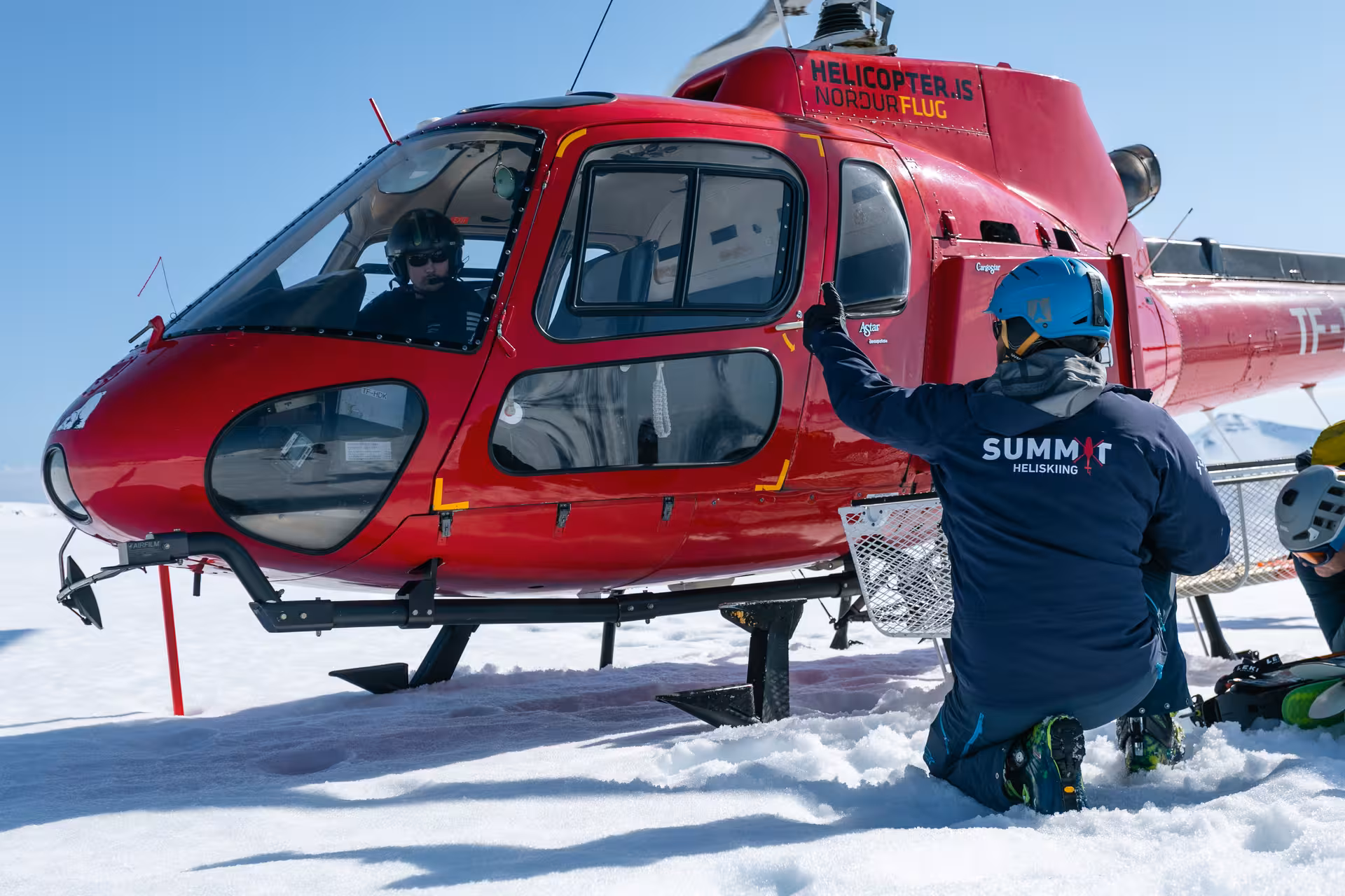 Red helicopter on snowy terrain with guide signaling for an exciting heli-skiing expedition.