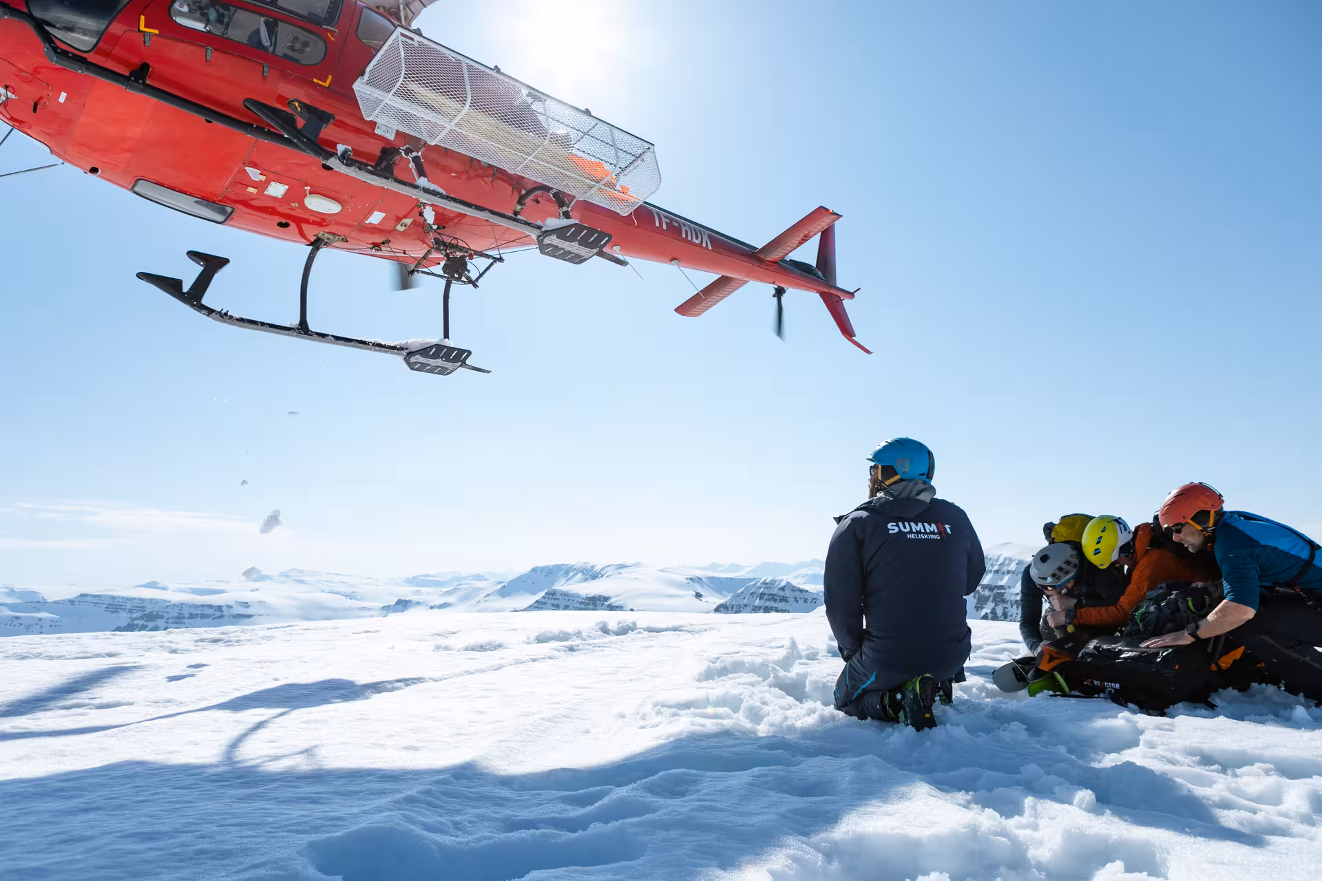 Red helicopter hovering over snow-capped mountain with skiers preparing gear during exhilarating heli-tour experience.