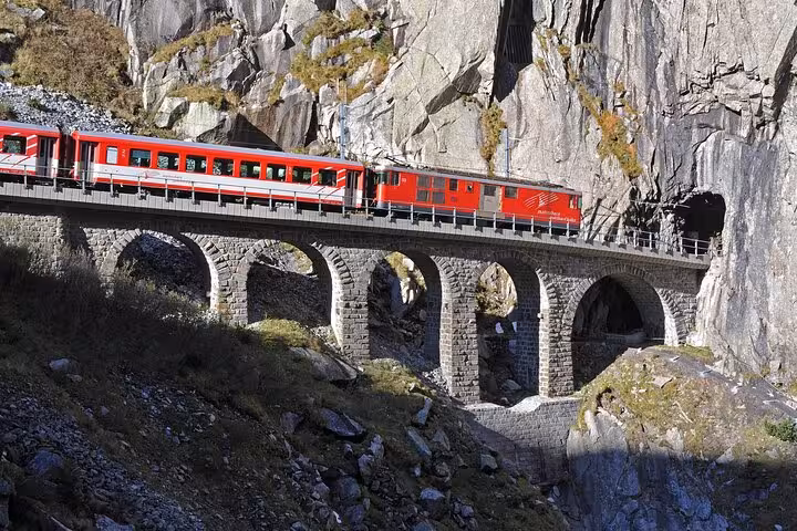 Red train crossing a stone viaduct amidst the rugged Swiss Alps landscape.