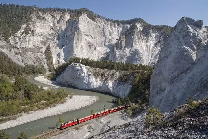 Scenic view of a red train winding through the dramatic cliffs and river valleys of the Swiss Alps.