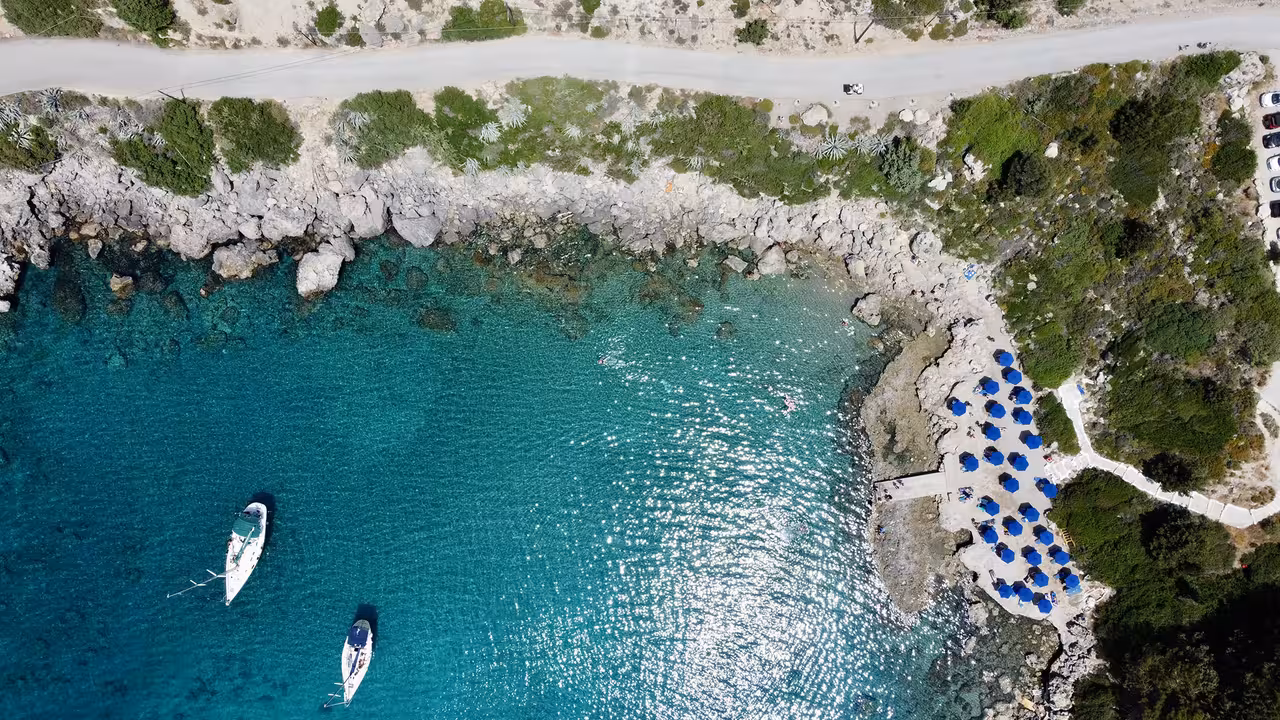 Top view of yachts anchored in crystal-clear waters next to a rocky coast lined with blue parasols on Regal 2700 tour.