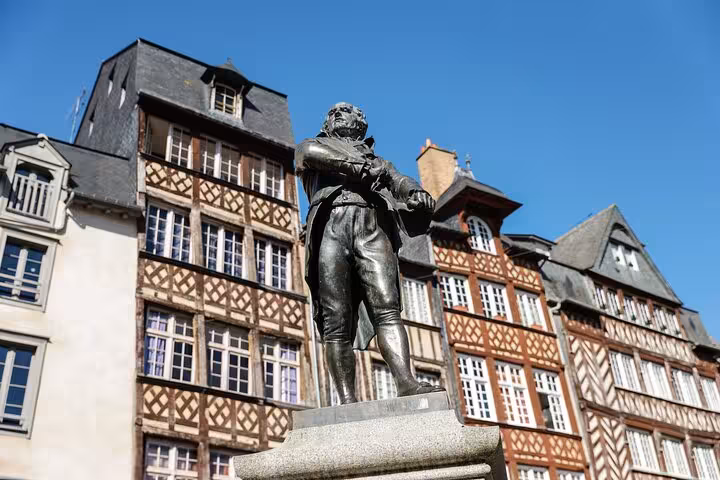 Statue of a historical figure in front of traditional half-timbered houses in Rennes, France, on a sunny day.