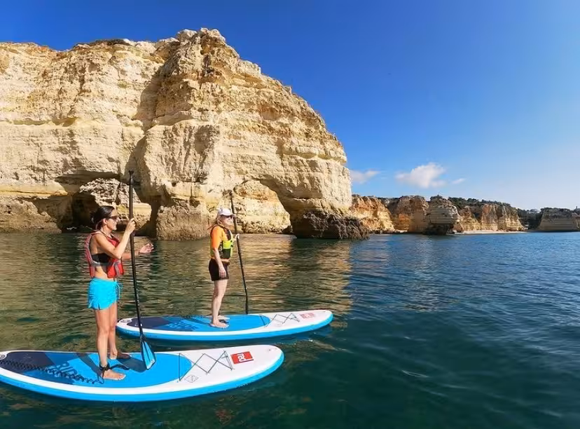 Two women on rented stand up paddleboards exploring calm turquoise waters and dramatic limestone cliffs in Algarve