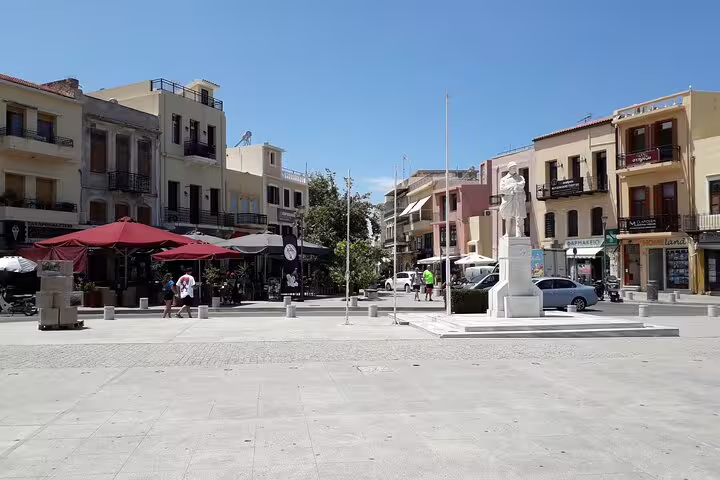 Bustling square in Rethymno Town with colorful buildings, outdoor cafes, and a central statue under a clear blue sky.