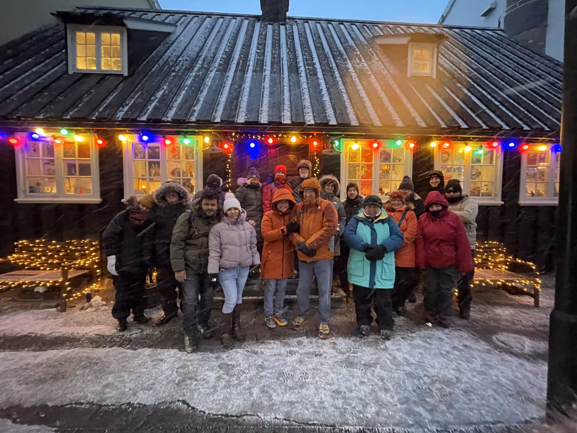Group of tourists in winter attire standing outside a cozy Reykjavík house adorned with colorful lights, ready for a cultural tour.