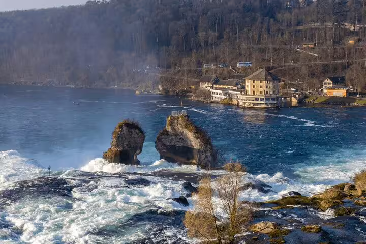 Scenic view of Rhine Falls with iconic rock formations, surrounded by lush greenery and historic architecture.