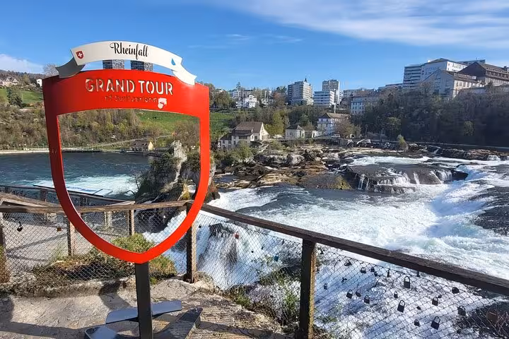 Scenic view of Rhine Falls with a Grand Tour sign, showcasing the majestic waterfall and surrounding buildings.