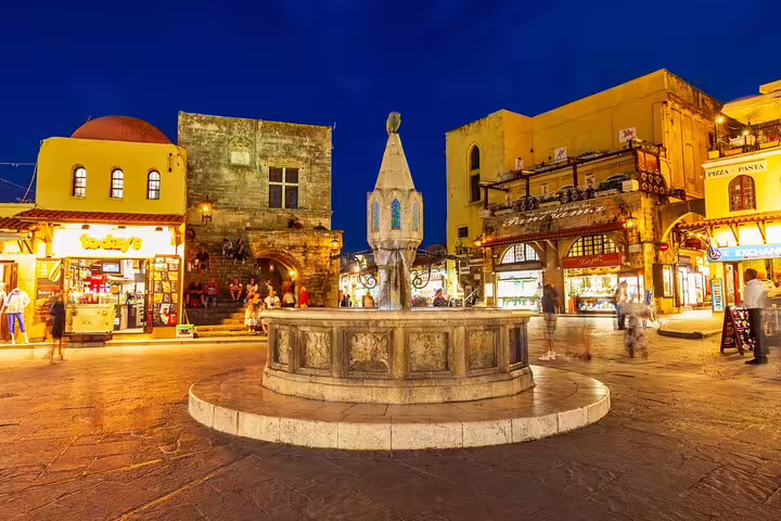Lively evening scene at Rhodes Old Town square, illuminated shops and historic fountain capturing local life ambiance.