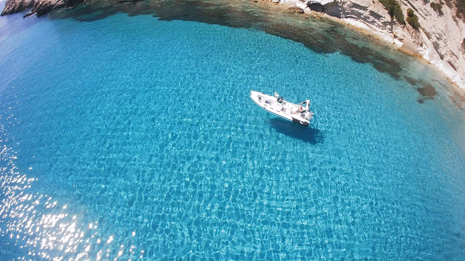 Aerial view of a RIB boat floating on crystal clear turquoise waters near rocky coastline in Carloforte, ideal for Sant'Antioco tours.