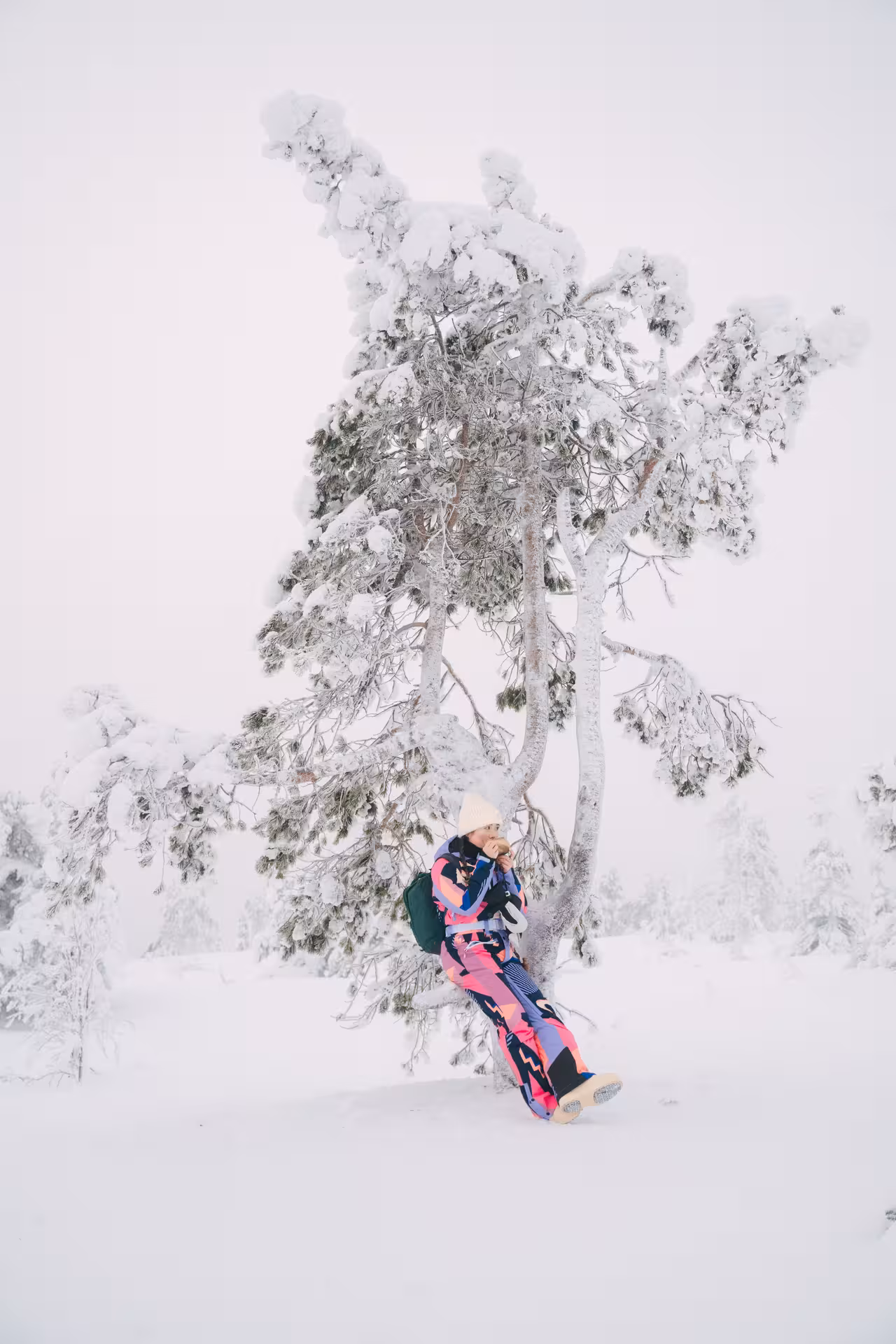 Traveler in vibrant snowsuit enjoying a break under a snow-laden tree in Riisitunturi's winter landscape.