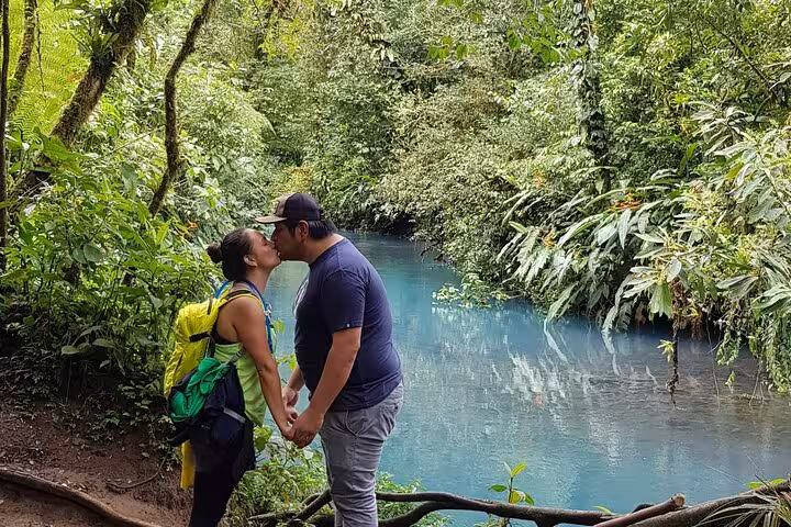 Couple enjoying a romantic moment by the vibrant blue waters of Rio Celeste amidst lush rainforest scenery.