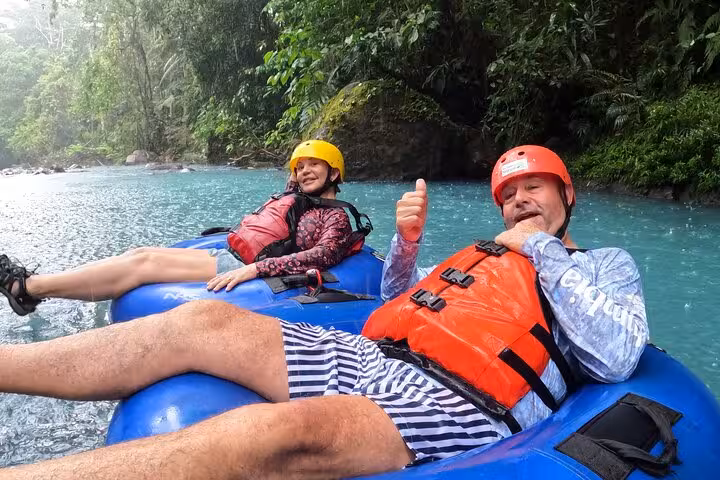 Two adventurers in helmets and life jackets tubing down the vibrant blue waters of Rio Celeste rainforest.