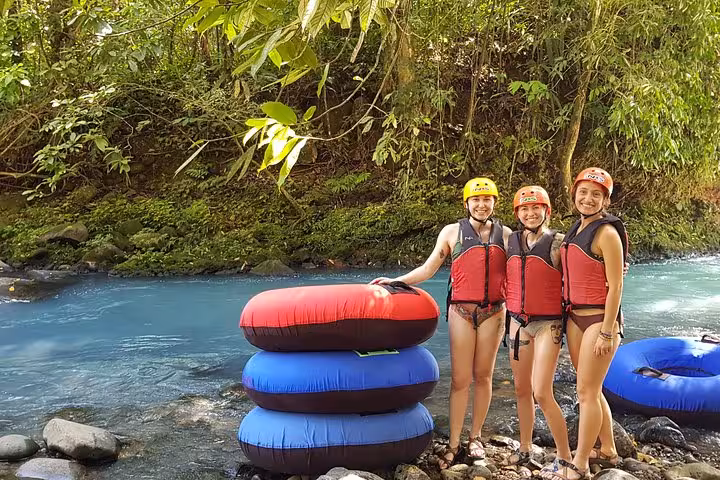 Adventurers in life jackets pose with inner tubes by the clear blue river, ready for tubing in Rio Celeste's lush surroundings.