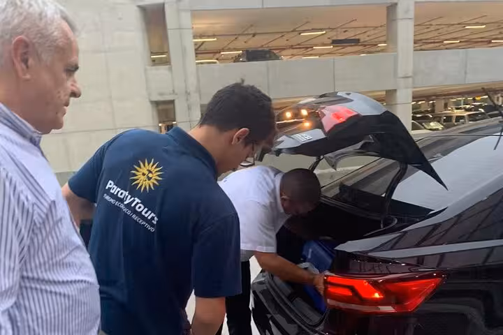 Travelers load luggage into a car trunk at a Rio de Janeiro airport, preparing for a shared shuttle to Angra dos Reis.
