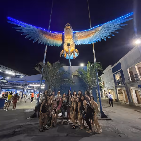 Group of people posing under a giant bird sculpture at Rio's samba school rehearsal, highlighting carnival excitement.