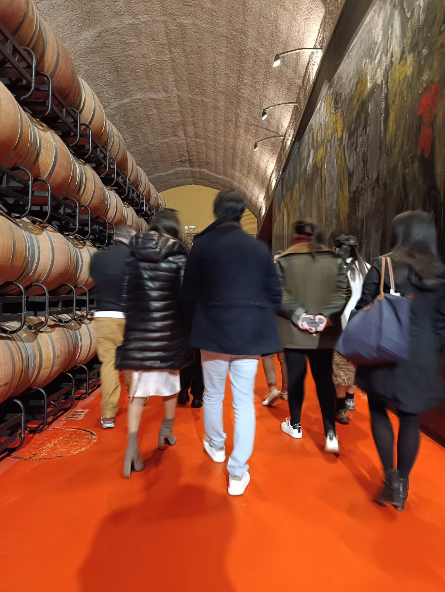 Tour group exploring Rioja wine cellar with rows of oak barrels on vibrant red flooring, highlighting wine aging process.