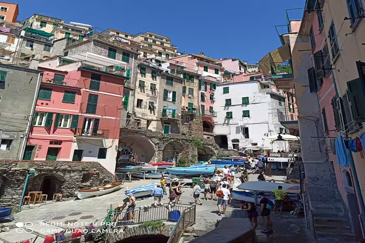 Tourists explore Riomaggiore harbor in Cinque Terre, surrounded by colorful cliffside houses and boats on a sunny Italy day