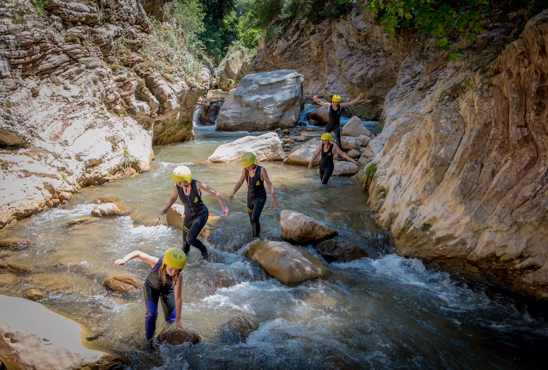 Group in helmets and wetsuits river trekking through Neda canyon, stepping on boulders in clear stream