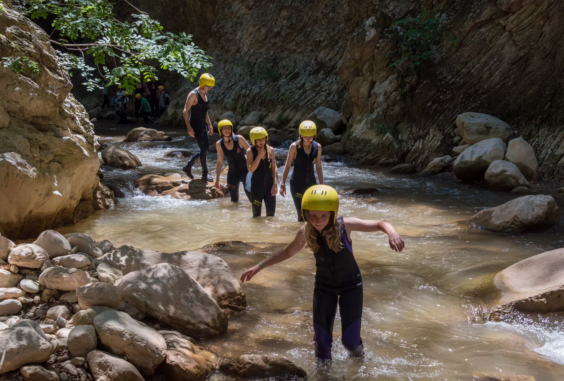River trekking group wading through shallow stream in Neda Gorge, guided canyon hike with helmets and wetsuits