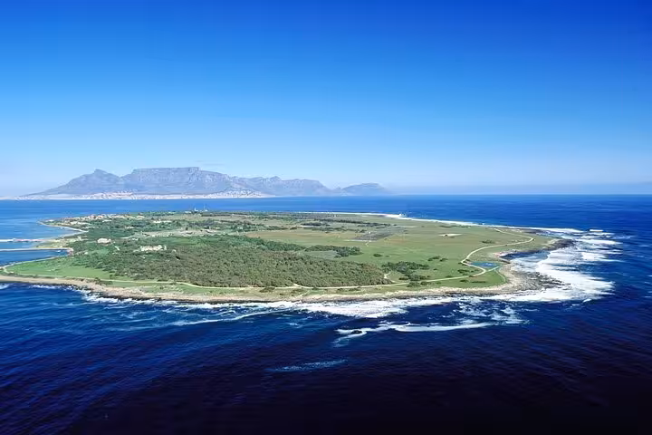 Aerial view of Robben Island surrounded by blue ocean, with Table Mountain visible in the distance.