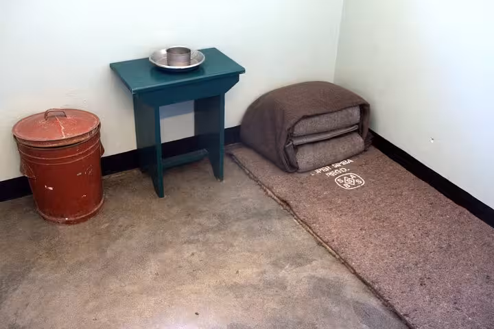 Interior of a sparse cell on Robben Island, showing a bedroll, small table with a metal dish, and a red bucket.