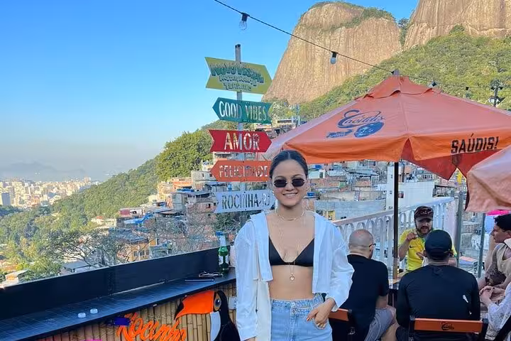 A visitor enjoys a rooftop view in Rocinha with colorful signs and scenic mountains on a sunny day.