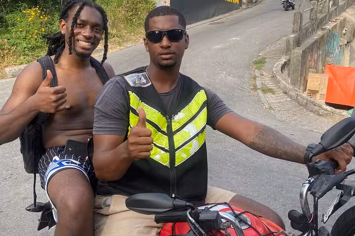 Two men on a motorcycle, one in a reflective vest, giving thumbs up during a Rocinha favela tour in Rio de Janeiro.