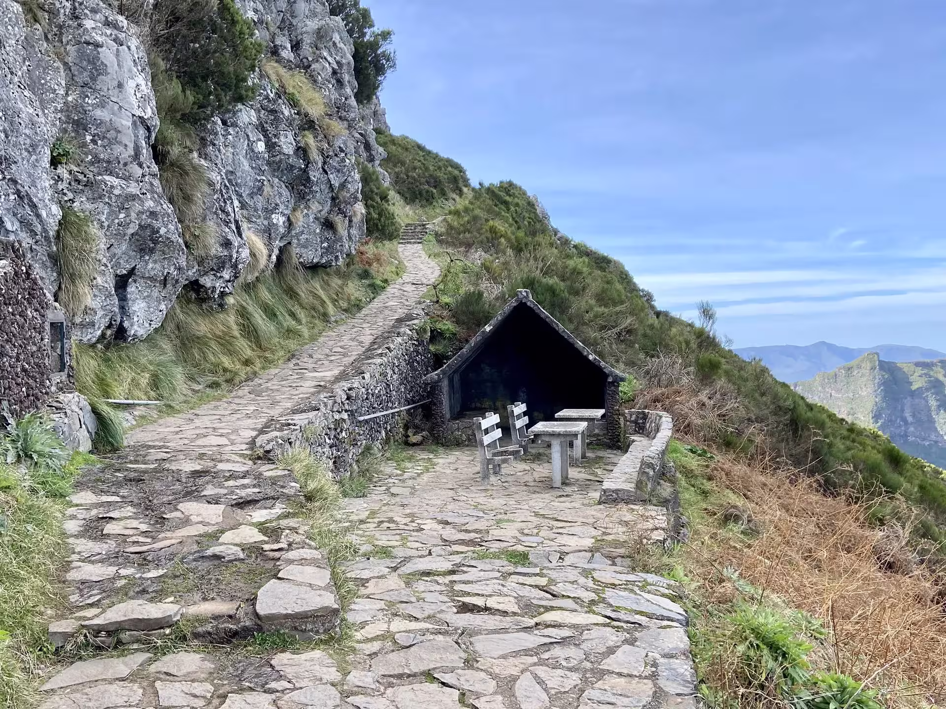 Scenic hiking path on rocky mountain with benches, offering panoramic views of lush landscapes and blue skies.