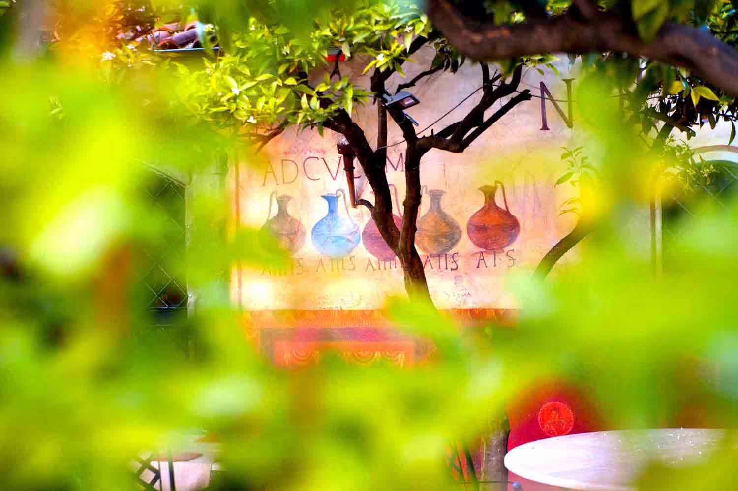 Lush garden corner of a Roman caupona with frescoed amphorae wall, leafy trees and outdoor New Year’s Eve seating
