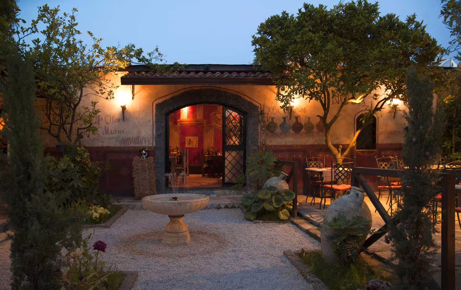 Evening view of illuminated Roman caupona courtyard with fountain, trees and outdoor tables for New Year’s Eve celebration