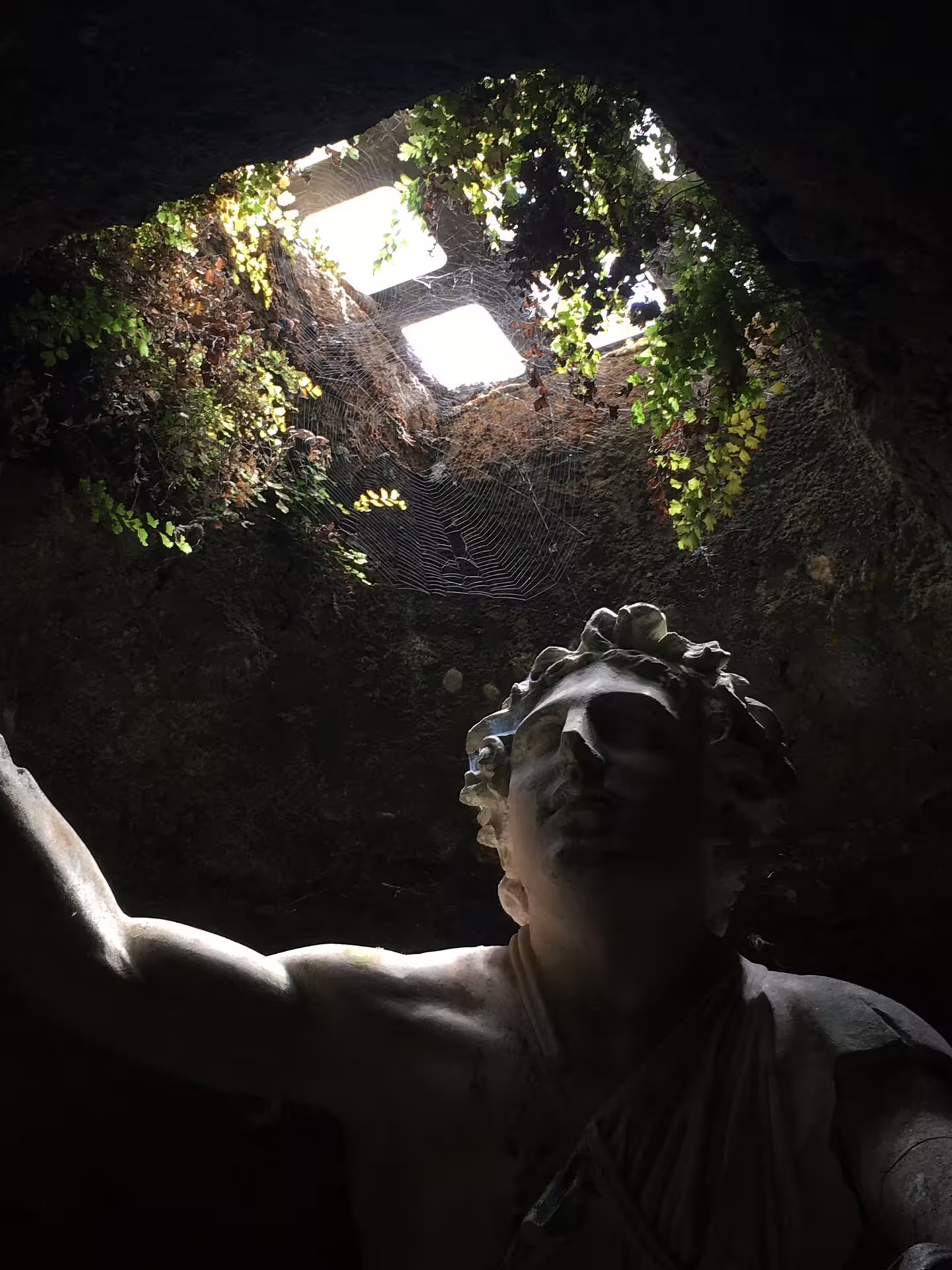 Ancient Roman statue lit from a cave skylight with cobwebs and ivy, evoking eerie legends on a Ghosts of Rome tour