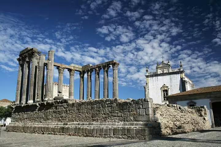 The Roman Temple of Évora, an ancient UNESCO World Heritage site, stands beside a historic church under a cloudy sky.